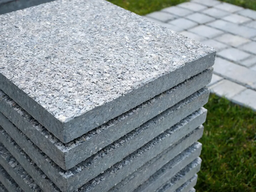 Betonowa płyta chodnikowa na stosie podobnych, z widoczną fakturą i krawędziami, na tle ułożonego chodnika.